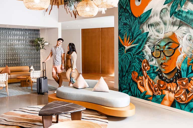 Modern hotel lobby with tropical mural art, woven pendant lights, and guests with luggage