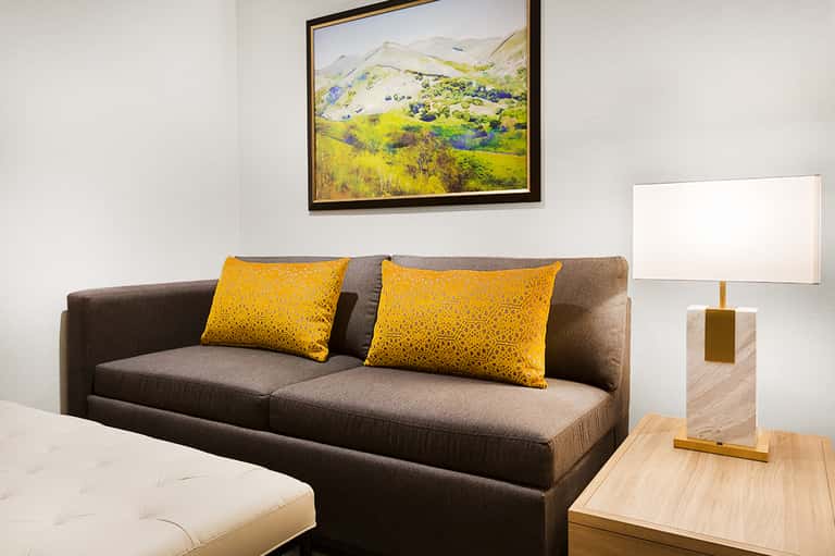 Modern hotel suite living room with gray sofa, yellow pillows, and landscape artwork