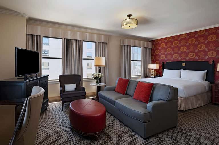 Luxurious hotel suite with red patterned walls, gray sofa, white bedding, and city views