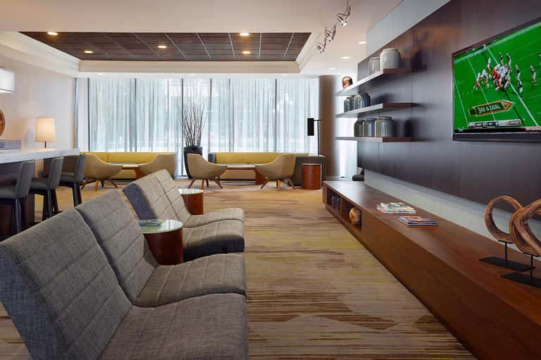 Modern hotel lounge with yellow accent chairs, TV, shelving, and floor-to-ceiling windows