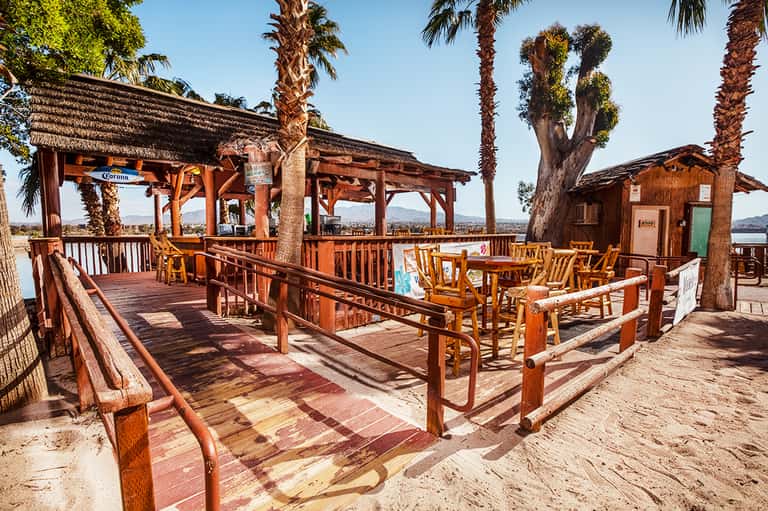 Rustic waterfront bar deck with wooden structures, palm trees, and lake or waterway views