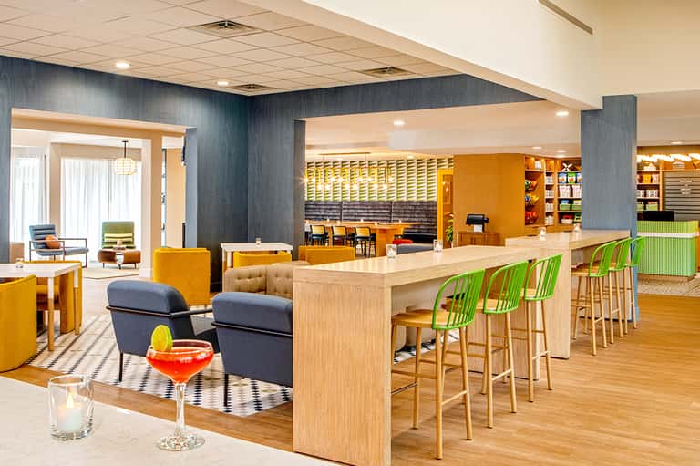 Contemporary hotel lobby bar with orange and green accents, wooden furnishings, and open layout