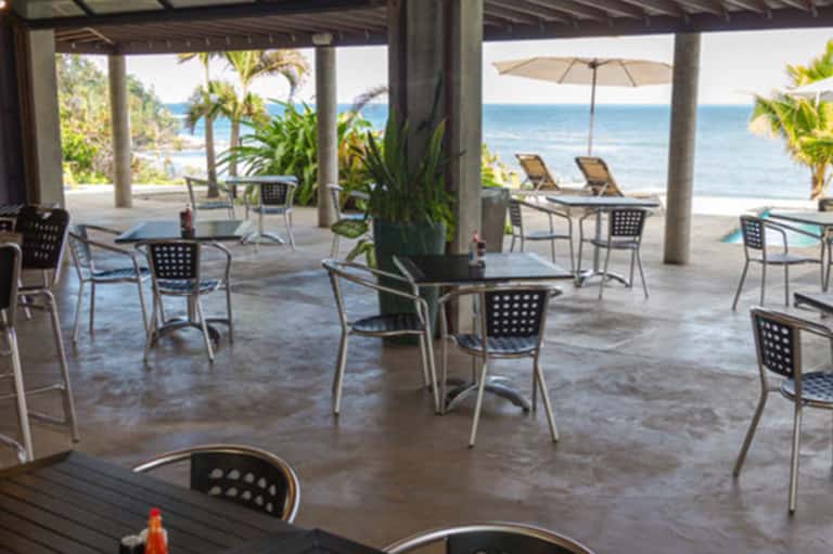 Beachfront dining area with ocean views, covered patio, tables with dark chairs, palm trees, and sandy beach