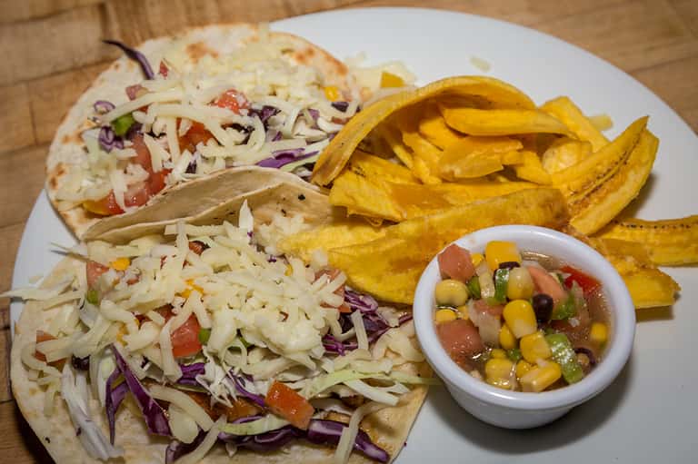 Plate of tacos with shredded cabbage, golden plantain chips, and corn salsa bowl