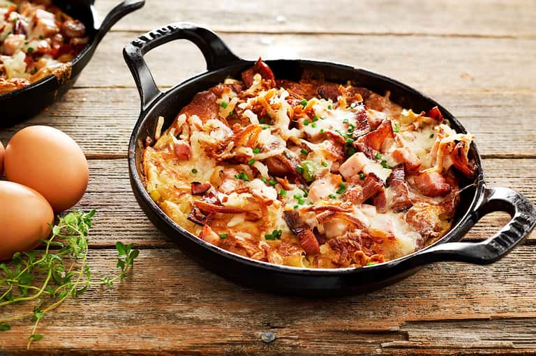 Skillet breakfast dish with eggs, bacon, and cheese on rustic wooden table with fresh herbs