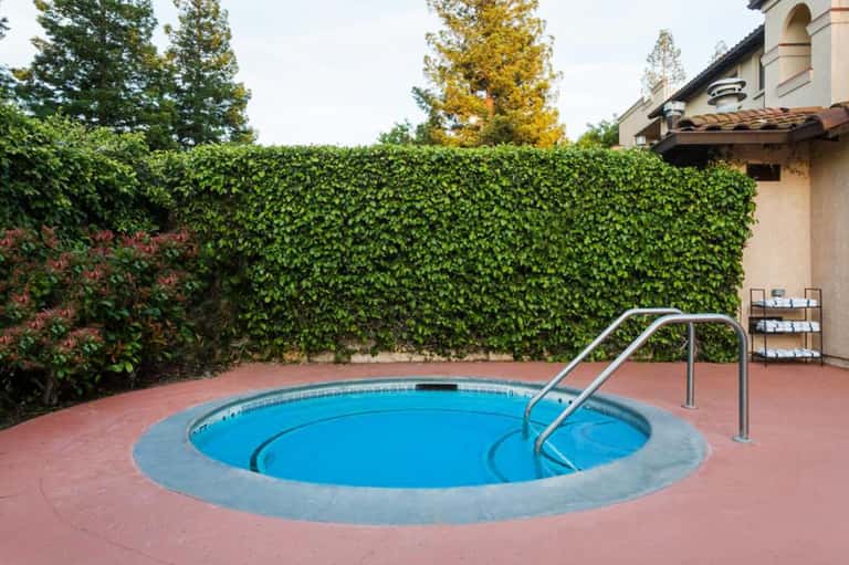 Round hot tub spa surrounded by brick patio, green ivy hedge wall, and metal railings in private resort courtyard
