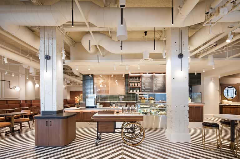 Modern hotel lobby and café with industrial ceiling, striped flooring, wooden furniture, and contemporary design elements