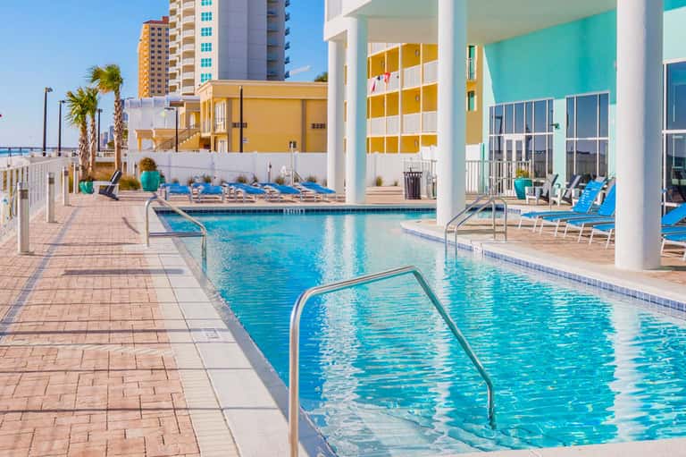 Bright turquoise pools with lounge chairs, colorful modern buildings, and palm trees at a beachfront resort