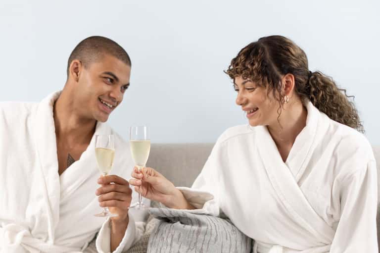 Couple in white robes clinking champagne glasses together on couch against light background