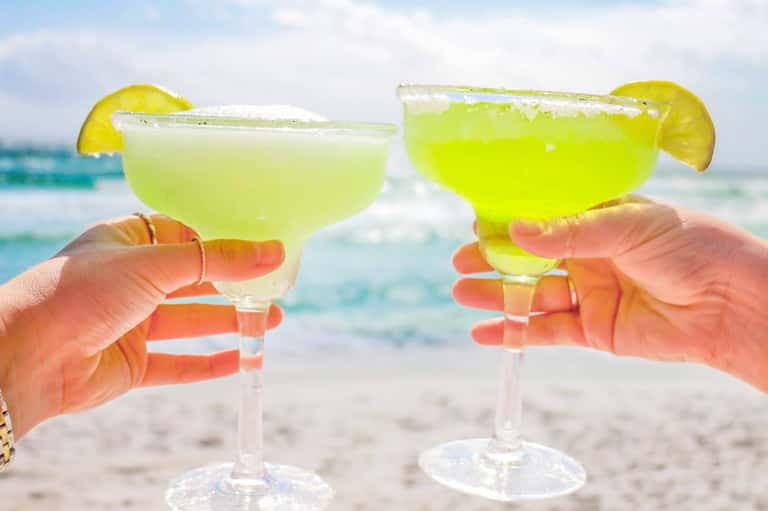 Two margaritas with lime slices and salted rims held up at a beach, ocean in background