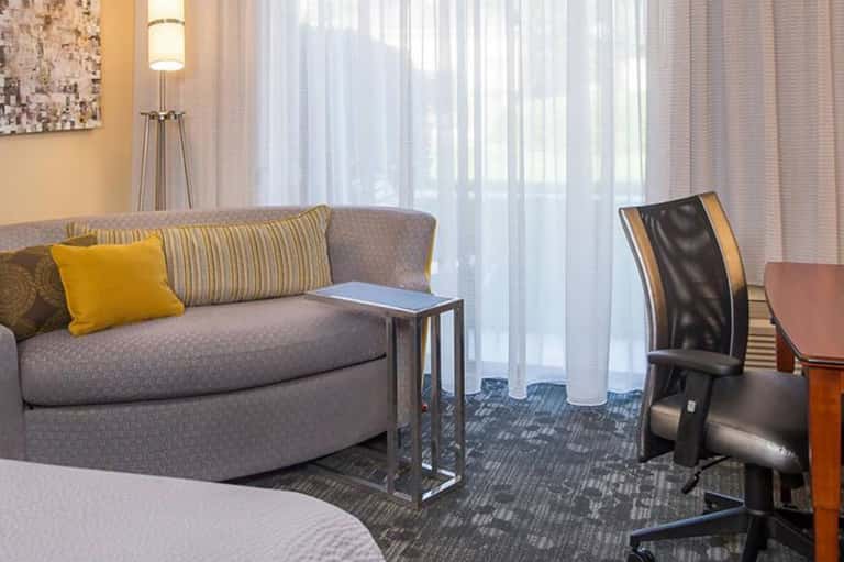 Hotel suite with curved gray sofa, yellow accent pillow, work desk with black chair, and sheer curtains