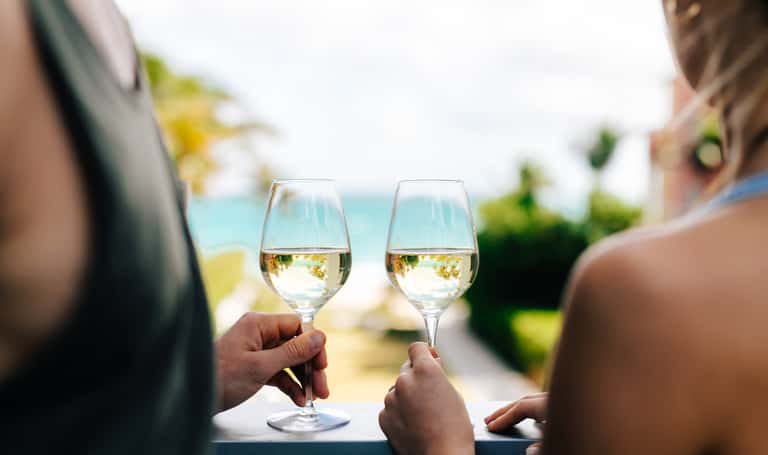 Two people toasting with white wine glasses overlooking a blue ocean coastline