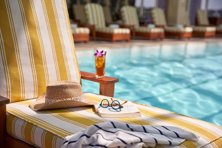 Poolside lounge chair with yellow striped fabric, straw hat, sunglasses, and cocktail with flower garnish