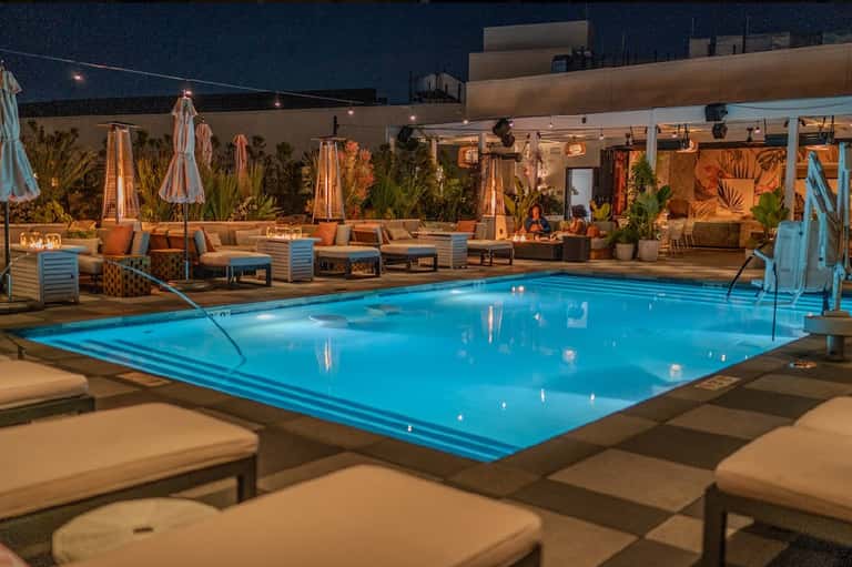 Nighttime resort pool illuminated in blue with fire pits, lounge seating, and striped umbrellas