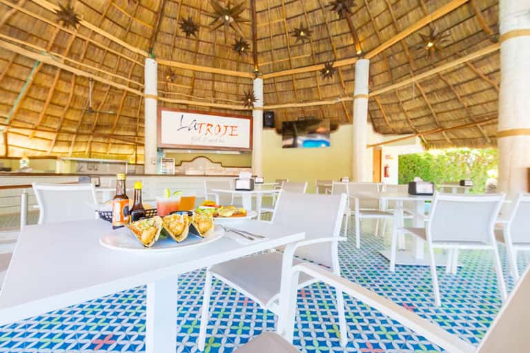 Open-air restaurant under thatched roof with white furniture and tacos on table