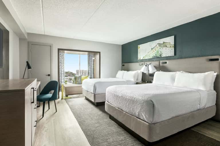 Hotel guest room with two queen beds, teal accent wall, desk, and balcony with city views