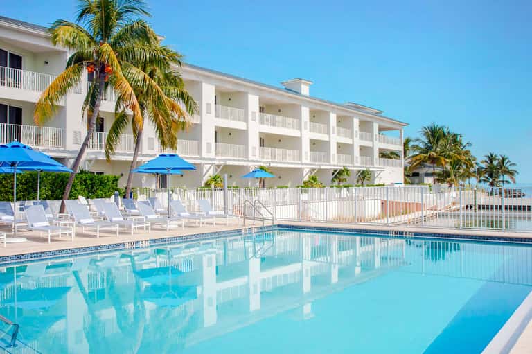 Waterfront resort with white building, blue umbrellas, lounge chairs, and crystal-clear pool overlooking the ocean