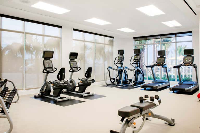 Bright fitness center with cardio equipment, exercise bikes, treadmills, and large windows overlooking palm trees