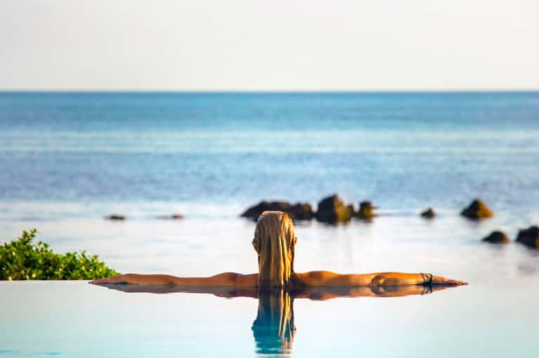 Woman in infinity pool with arms outstretched overlooking ocean and rocky coastline at sunset