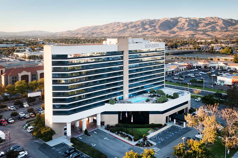 Modern glass hotel building with pool on rooftop, mountains and water visible in background landscape