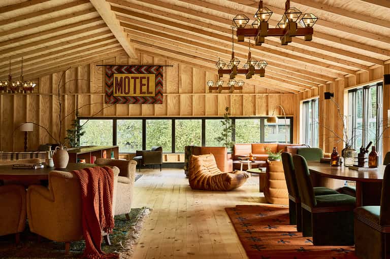 Retro motel-style lobby with wood beams, vintage furniture, and large windows overlooking forest