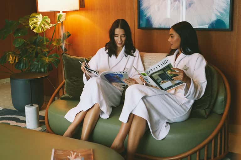 Two women in white spa robes relaxing on a green sofa reading magazines