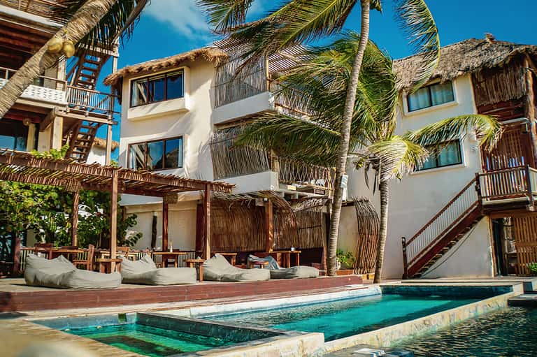 Beachfront resort buildings with thatched-roof structures, white walls, turquoise pool, and palm trees