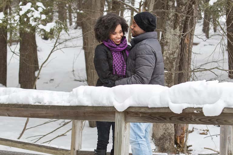 Couple in winter clothing embracing on snowy wooden fence with frozen lake and bare trees behind