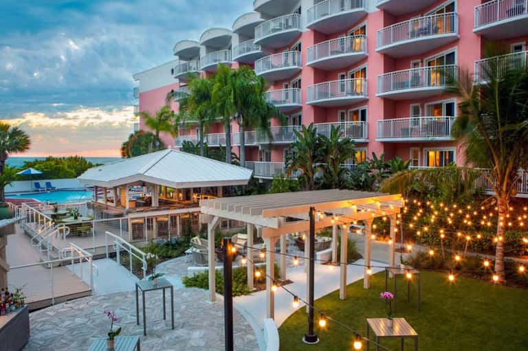 Waterfront resort at dusk with pink building, illuminated pergola, pool area, and palm-lined grounds