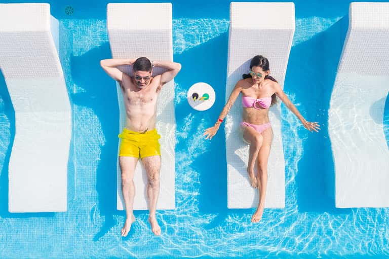 Two people floating on white loungers in bright blue pool with mirrored reflections below