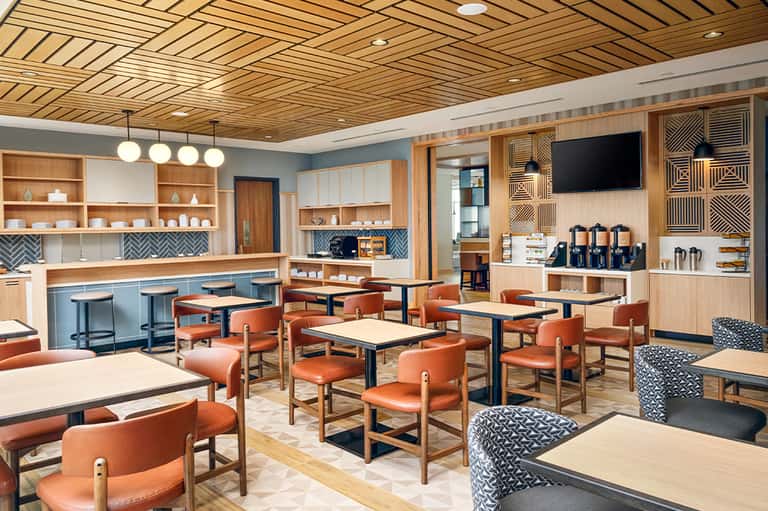 Modern café lounge with wood ceiling, orange chairs, blue accents, and beverage station