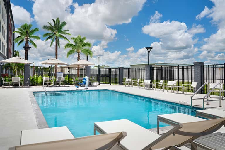 Resort pool with white lounge chairs, umbrellas, palm trees, and black fence railing
