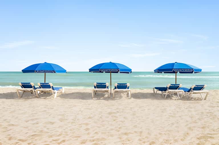 Beach scene with three blue umbrellas, white lounge chairs, turquoise ocean, and sandy shore