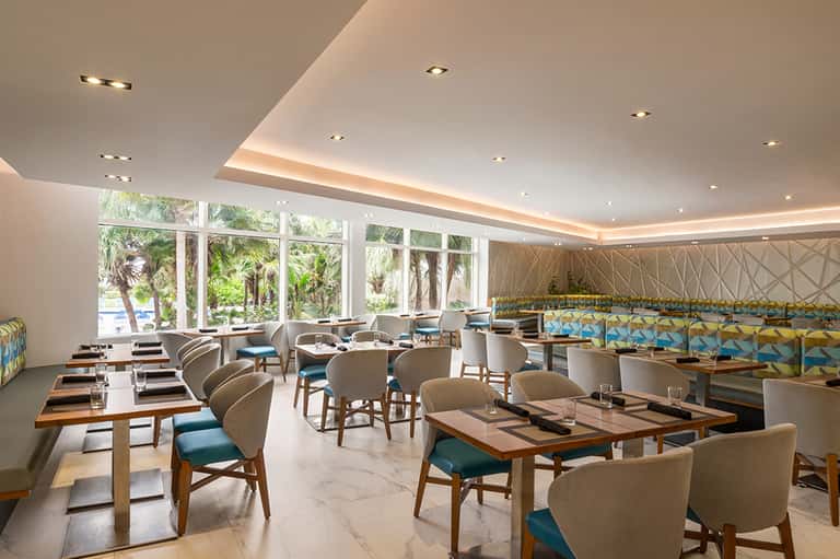 Contemporary restaurant with large windows, palm tree views, wooden tables, and beige upholstered chairs