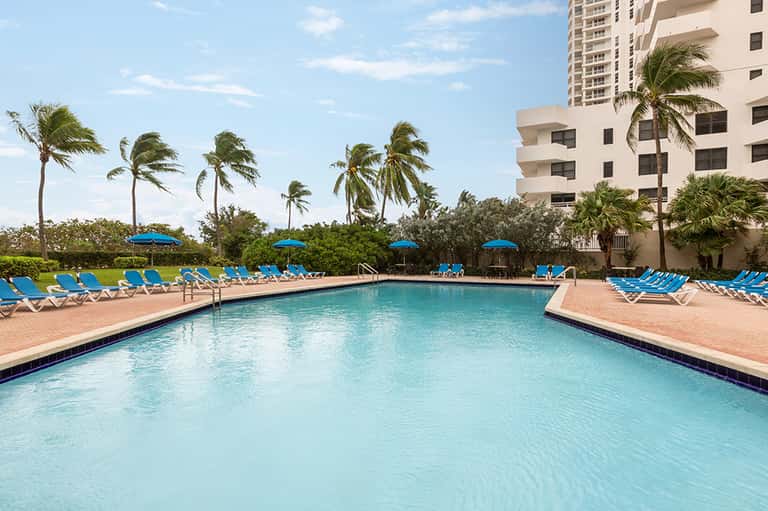 Large rectangular resort pool with blue lounge chairs and umbrellas, flanked by palm trees and beige buildings