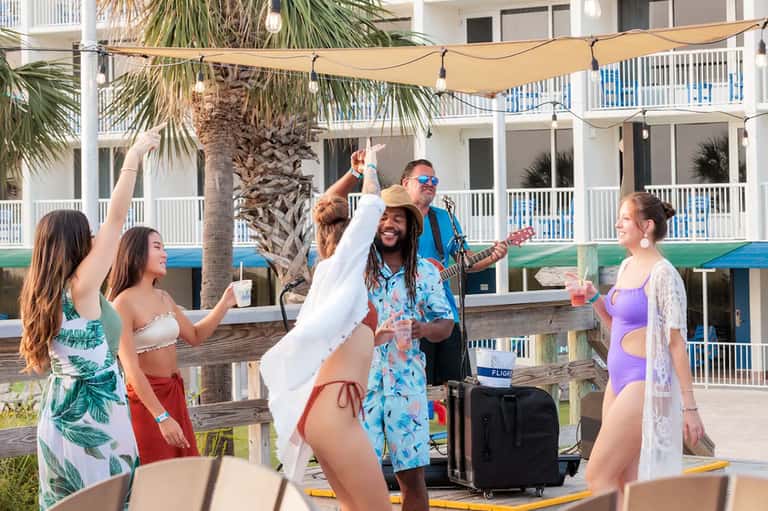 Beach musicians entertaining guests under shaded area with palm trees and resort building