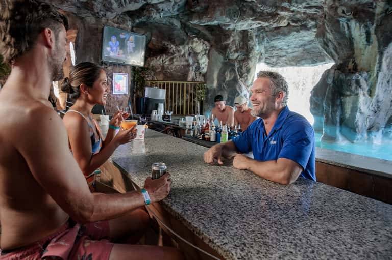 Beach bar built into cave with customers enjoying drinks, water view backdrop, and casual resort setting