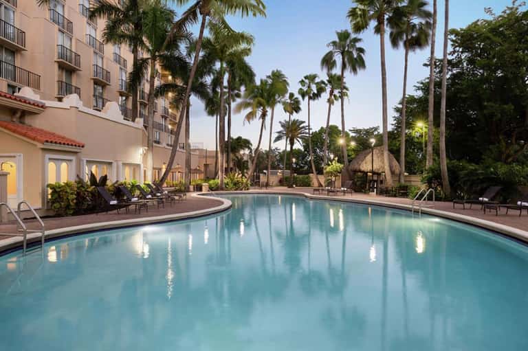Resort pool at dusk with manicured landscape, palm trees, and adjacent hotel building