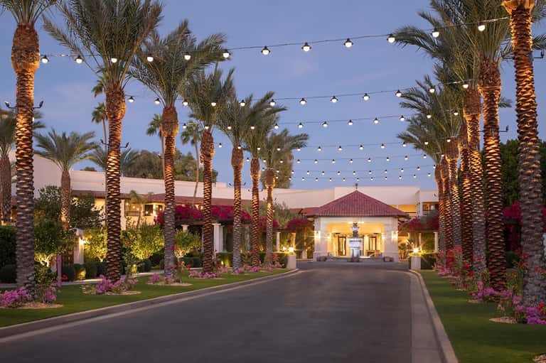 Palm-lined driveway lit by string lights and uplighting, leading to illuminated Spanish-style villa entrance at dusk