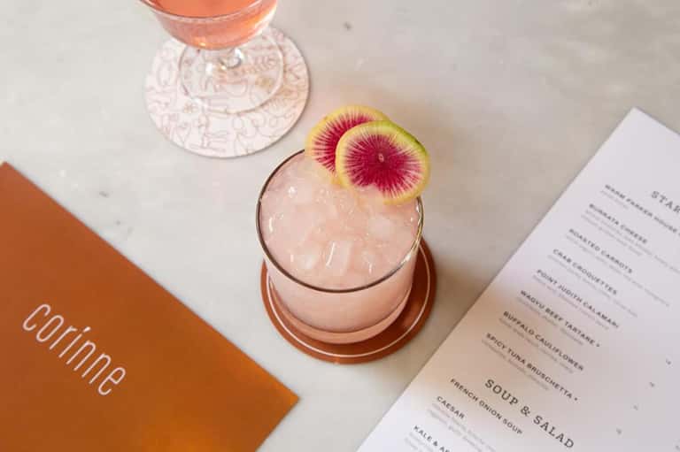 Elegant pink cocktail with watermelon slice garnish next to menu and place setting on white marble table