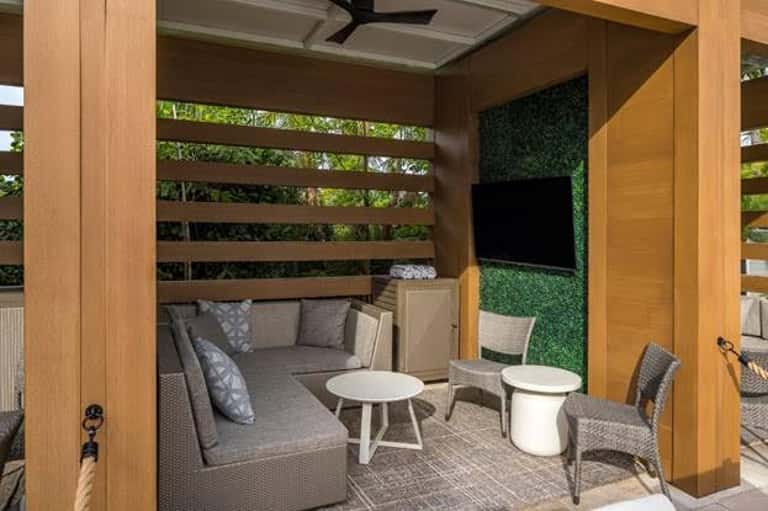 Private cabana with wooden frame, green wall, comfortable seating, white side tables, and TV mount