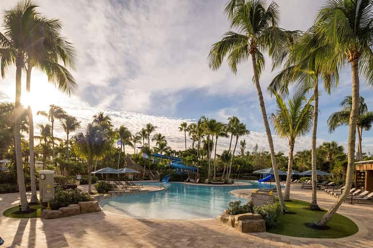 Resort lagoon pool with sandy beach entry, palm trees, blue waterslide, and tropical landscaping