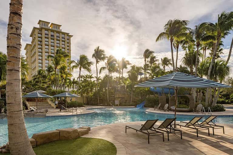 Resort pool with blue umbrellas, lounge chairs, palm trees, and hotel building backdrop