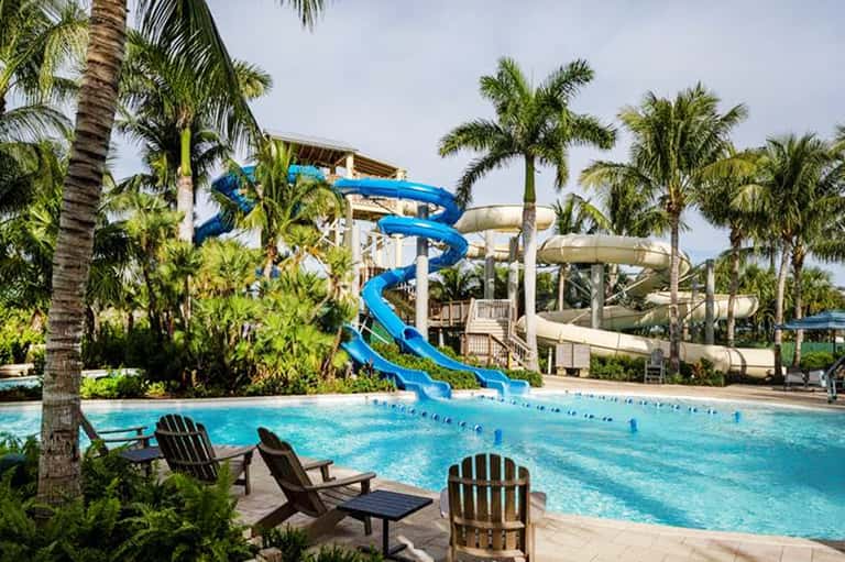 Waterslide pool complex with blue spiral slide, palm trees, and Adirondack lounge chairs