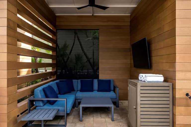 Private patio cabana with blue sectional sofa, wood walls, window slats, and TV mounted on wall
