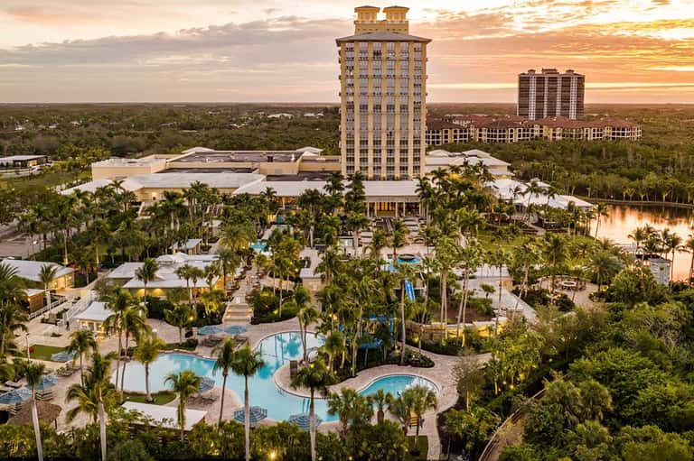Aerial resort view at sunset showing tall tower, pools, palm trees, and surrounding natural landscape