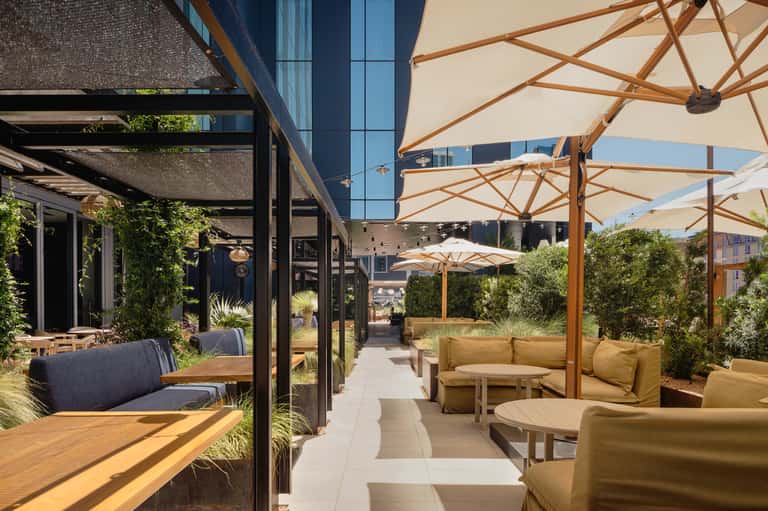 Upscale rooftop terrace with tan umbrellas, modern seating, lush plants, and city views in background