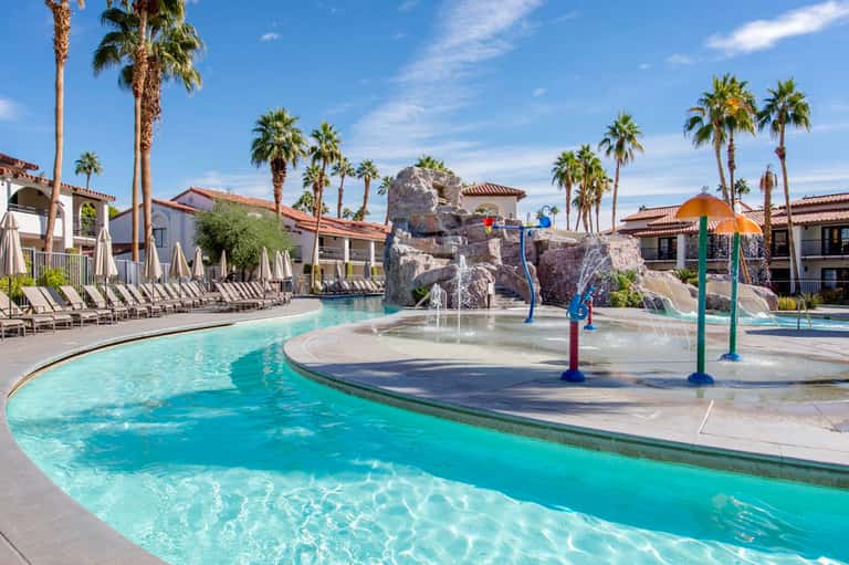 Resort pool with rock waterfall feature, splash pad, and palm trees with Spanish colonial architecture