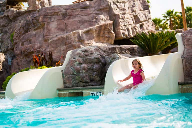 Child sliding down water slide into turquoise pool with rocky landscape backdrop