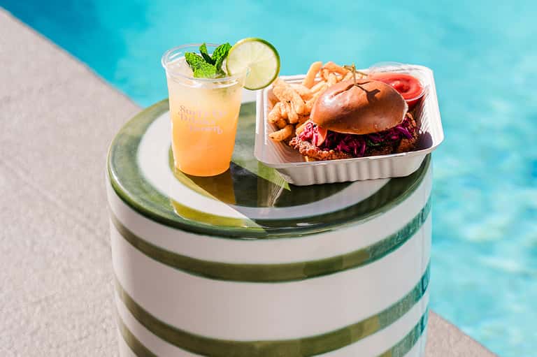 Poolside meal with iced beverage and chicken sandwich served on striped table at resort with turquoise water